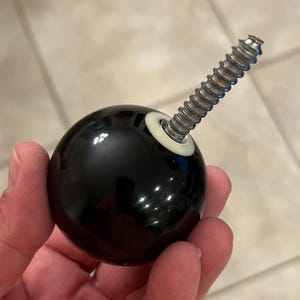 Puede incluir: Un pomo de armario esférico negro con un tornillo plateado que se extiende desde el centro. El pomo tiene un acabado brillante y una arandela blanca. El tornillo es de metal y tiene un diseño roscado. El pomo se sostiene en una mano.