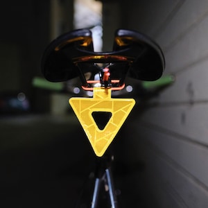 Może przedstawiać: Czarne siodełko rowerowe z ż&oacute;łtym tr&oacute;jkątnym odblaskiem. Odbłyśnik ma geometryczny wz&oacute;r i jest przymocowany do sztycy podsiodłowej. Tło jest ciemne i rozmyte.