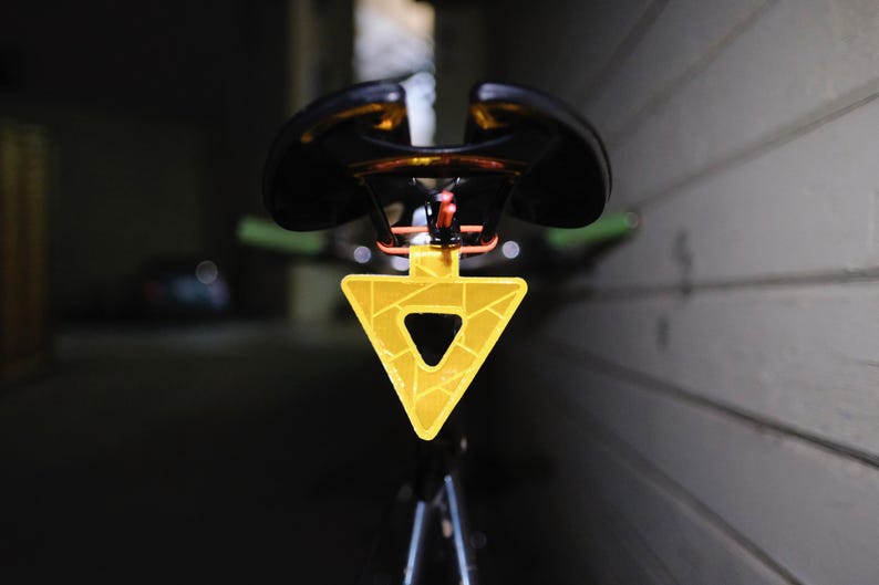 Może przedstawiać: Czarne siodełko rowerowe z ż&oacute;łtym tr&oacute;jkątnym odblaskiem. Odbłyśnik ma geometryczny wz&oacute;r i jest przymocowany do sztycy podsiodłowej. Tło jest ciemne i rozmyte.