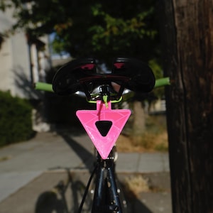 Może przedstawiać: Siodło rowerowe z czarnym siedziskiem i zieloną kierownicą. Jasnor&oacute;żowy, tr&oacute;jkątny odblask jest przymocowany pod siodełkiem. Rower jest ustawiony przed drzewem i budynkiem.