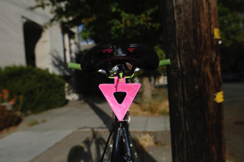 Może przedstawiać: Siodło rowerowe z czarnym siedziskiem i zieloną kierownicą. Jasnor&oacute;żowy, tr&oacute;jkątny odblask jest przymocowany pod siodełkiem. Rower jest ustawiony przed drzewem i budynkiem.