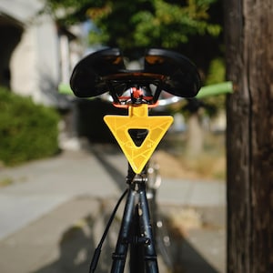 Może przedstawiać: Czarne siodełko rowerowe z jasnoż&oacute;łtym tr&oacute;jkątnym odblaskiem. Odblask ma geometryczny wz&oacute;r i centralne wycięcie. Kierownica ma zielone uchwyty.