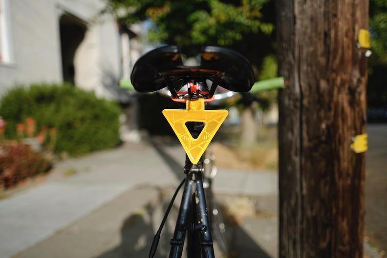 Może przedstawiać: Czarne siodełko rowerowe z jasnoż&oacute;łtym tr&oacute;jkątnym odblaskiem. Odblask ma geometryczny wz&oacute;r i centralne wycięcie. Kierownica ma zielone uchwyty.