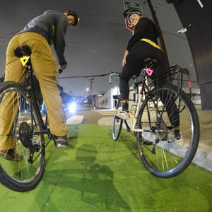 Może przedstawiać: Dwie osoby jadące na rowerach po zielonym pasie rowerowym w nocy. Rowery mają czarne ramy i koła. Jeźdźcy mają na sobie kaski i ubrania codzienne. Scena jest oświetlona przez latarnie uliczne.