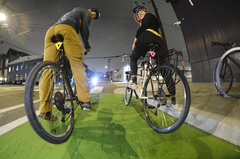 Może przedstawiać: Dwie osoby jadące na rowerach po zielonym pasie rowerowym w nocy. Rowery mają czarne ramy i koła. Jeźdźcy mają na sobie kaski i ubrania codzienne. Scena jest oświetlona przez latarnie uliczne.