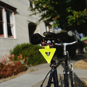 Może przedstawiać: Czarny rower z odblaskowym ż&oacute;łtym tr&oacute;jkątem przymocowanym do sztycy podsiodłowej. Rower ma czarne siodełko, srebrną kierownicę i zielone uchwyty kierownicy. Tło obejmuje budynek i liście.