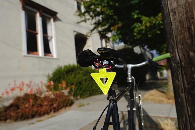 Może przedstawiać: Czarny rower z odblaskowym ż&oacute;łtym tr&oacute;jkątem przymocowanym do sztycy podsiodłowej. Rower ma czarne siodełko, srebrną kierownicę i zielone uchwyty kierownicy. Tło obejmuje budynek i liście.
