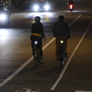 Może przedstawiać: Dw&oacute;ch rowerzyst&oacute;w jedzie rowerami nocą po drodze ze ścieżką rowerową. Rowerzyści mają na sobie ciemne ubrania i odblaskowe tr&oacute;jkąty na plecach. Reflektory pojazd&oacute;w oświetlają drogę.