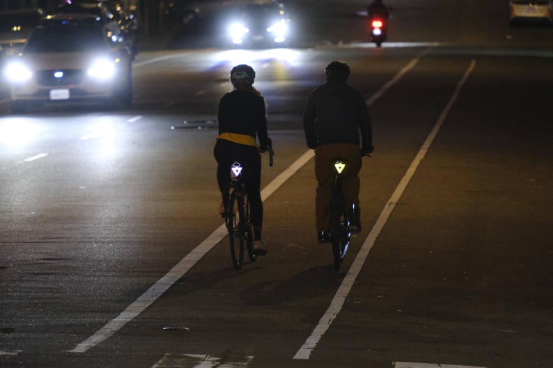 Może przedstawiać: Dw&oacute;ch rowerzyst&oacute;w jedzie rowerami nocą po drodze ze ścieżką rowerową. Rowerzyści mają na sobie ciemne ubrania i odblaskowe tr&oacute;jkąty na plecach. Reflektory pojazd&oacute;w oświetlają drogę.