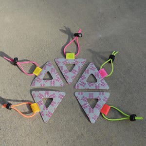 Może przedstawiać: Sześć szarych tr&oacute;jkątnych obiekt&oacute;w z otworem pośrodku, każdy z innym kolorowym sznurkiem i przełącznikiem. Tr&oacute;jkąty mają powtarzający się wz&oacute;r słowa "VISTO" w kolorze r&oacute;żowym. Sznurki są r&oacute;żowe, pomarańczowe i zielone.