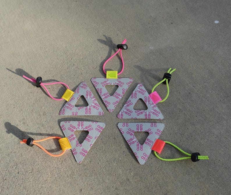 Może przedstawiać: Sześć szarych tr&oacute;jkątnych obiekt&oacute;w z otworem pośrodku, każdy z innym kolorowym sznurkiem i przełącznikiem. Tr&oacute;jkąty mają powtarzający się wz&oacute;r słowa "VISTO" w kolorze r&oacute;żowym. Sznurki są r&oacute;żowe, pomarańczowe i zielone.