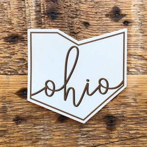 Könnte beinhalten: Ein weißes Holzschild in Form von Ohio mit braunem Rand und dem Wort "ohio" in Schreibschrift. Das Schild befindet sich vor einem Holzgrund.