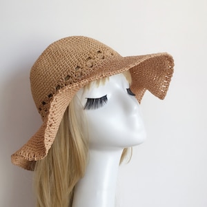 May include: A light brown crocheted sun hat with a wide brim and a delicate crocheted design. The hat is worn on a mannequin with long blonde hair.