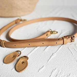 May include: A light brown leather strap with two metal clasps and two leather tabs. The strap is on a white surface.