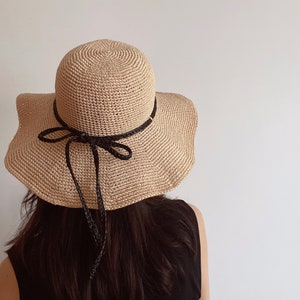 Puede incluir: Un sombrero de paja de ala ancha con una cinta negra atada en un lazo. El sombrero está hecho de material de crochet y tiene un color beige natural.