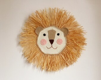 May include: A crocheted lion head wall hanging with a beige mane and a white face with brown eyes and pink cheeks.