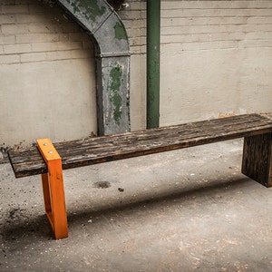 Puede incluir: Un banco de madera r&uacute;stico con un acabado marr&oacute;n oscuro y patas de metal naranja. El banco est&aacute; hecho de madera recuperada y tiene un dise&ntilde;o simple e industrial.