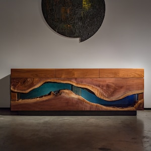 May include: A modern wooden sideboard with a live edge design. The natural wood grain is complemented by a vibrant blue epoxy river that runs through the center. The cabinet has three drawers and a dark metal base.