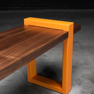 K&ouml;nnte beinhalten: Eine moderne Holzbank mit rechteckigem Design. Die Bank hat eine dunkelbraune Holzplatte und leuchtend orangefarbene Metallbeine. Die Beine bilden eine quadratische Form und bilden einen auff&auml;lligen Kontrast zum Holz.