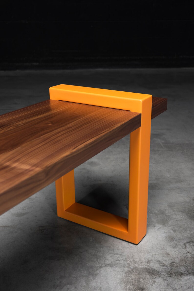 K&ouml;nnte beinhalten: Eine moderne Holzbank mit rechteckigem Design. Die Bank hat eine dunkelbraune Holzplatte und leuchtend orangefarbene Metallbeine. Die Beine bilden eine quadratische Form und bilden einen auff&auml;lligen Kontrast zum Holz.