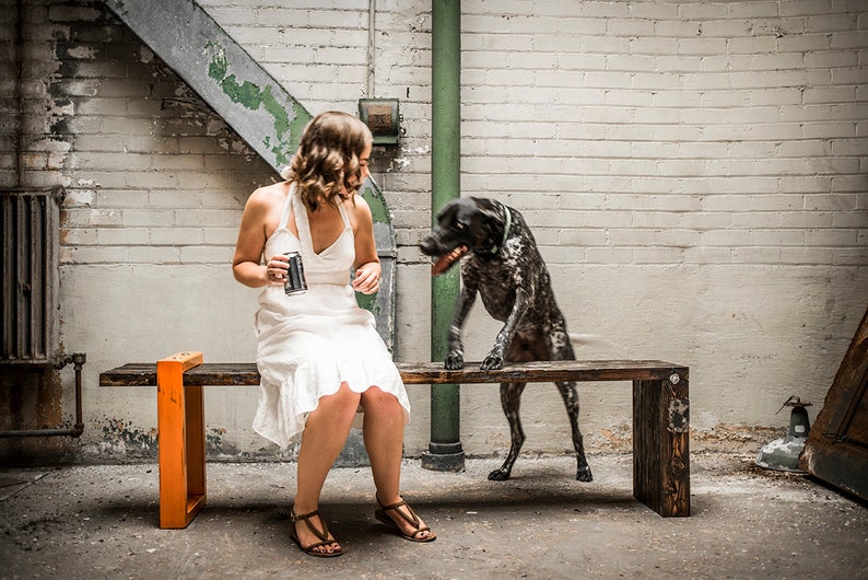 Puede incluir: Una mujer con un vestido blanco se sienta en un banco de madera con un marco de metal naranja. Un perro blanco y negro se para en el banco detr&aacute;s de ella.