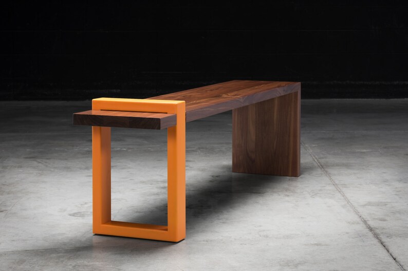 K&ouml;nnte beinhalten: Moderne Bank mit dunkler Holzsitze und einem kontrastierenden orangefarbenen St&uuml;tzrahmen. Die Bank hat eine rechteckige Sitzfl&auml;che und ein einzigartiges, geometrisches Design. Der orangefarbene Rahmen setzt Akzente zu den nat&uuml;rlichen Holzt&ouml;nen.