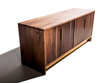May include: A rectangular wooden sideboard crafted from dark walnut wood featuring a slatted front design with vertical grooves. The cabinet includes two central doors with slim, vertical brass-colored metal handles and rests on a recessed brass base.