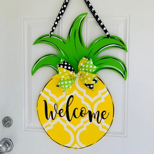May include: A yellow pineapple-shaped door hanger with a white and black geometric pattern and the word "welcome" written in black. The pineapple has green leaves and a yellow and green bow with white polka dots.