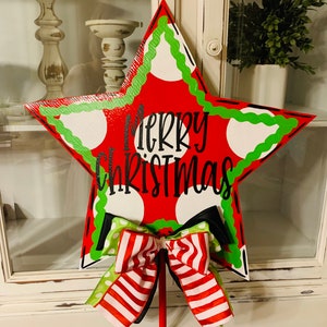 May include: A red star-shaped wooden sign with white polka dots and green trim. The sign says "Merry Christmas" in black lettering. A red, white, and green striped bow is attached to the sign.
