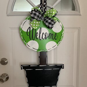 May include: A green and white polka dot welcome sign with a black and white checkered bow hanging on a white door. The sign is shaped like a tree and has a black pot.