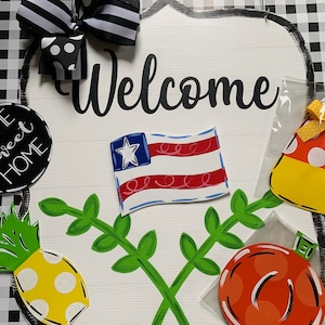 May include: A wooden welcome sign with a black and white checkered background. The sign features a red, white, and blue American flag, a green leafy vine, a yellow pineapple, and a red pumpkin. The sign also includes the word "Welcome" in black cursive lettering.