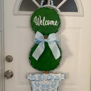 May include: A green welcome sign shaped like a topiary tree with a blue and white striped bow. The sign is hanging on a white door and has a blue and white patterned pot.