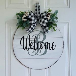 May include: A round wooden door hanger with a white background and gray and black buffalo plaid ribbon. The door hanger has a white and gray painted wood plank design and the word "Welcome" in black lettering. The door hanger is decorated with a gray monogram and greenery.