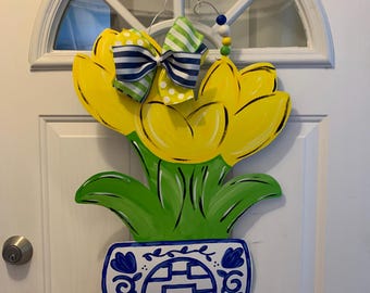 May include: A yellow tulip flower door hanger with green leaves and a blue and white patterned pot. The flower has a blue and white striped bow with white polka dots.