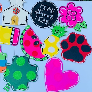 May include: A collection of colorful wooden signs with various designs, including a church, a watermelon slice, a pineapple, a heart, a shamrock, a paw print, a flower, and a sign that says "Home Sweet Home".
