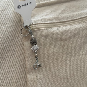 May include: A beaded keyring with a silver-tone metal ring and a decorative charm. The charm features a dark grey beaded ball, a clear crystal beaded ball, and silver-tone beads. The tag reads "BeadedKink" with a sunburst logo.