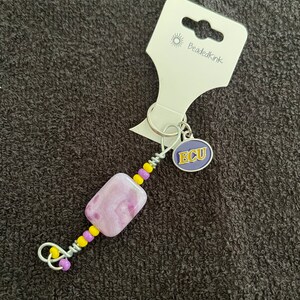 Peut inclure: Un porte-clés en perles avec une perle rectangulaire violette et blanche, des perles jaunes et violettes, et une boucle en métal argenté. Le porte-clés comporte un charm avec les lettres "ECU" sur fond violet. L'étiquette indique "BeadedKink".