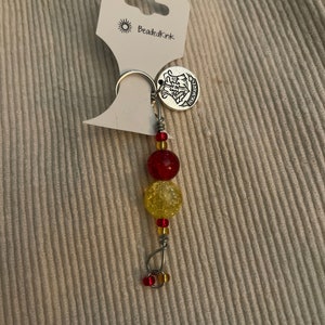 May include: A beaded keychain with red and yellow beads. The keychain has a silver-colored ring and a charm with the text "Hogwarts". The tag reads "Beadedkink".