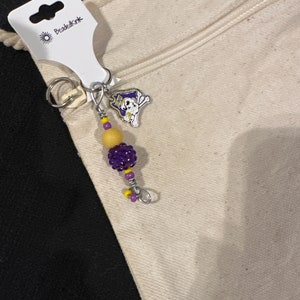 Peut inclure: Un porte-clés avec un charm en forme de chien de dessin animé dans un design violet et or. Le porte-clés a une breloque perlée avec des perles violettes et jaunes. L'étiquette indique "BeadedInk".
