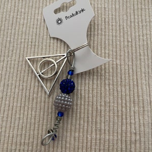 May include: A silver-toned charm with a triangle and circle design, attached to a beaded accessory. The accessory features blue and gray beads, with the text "BeadedKink" on the attached tag.