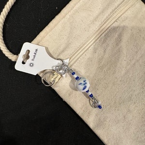 Könnte beinhalten: Ein Schlüsselanhänger mit Perlen mit einer weißen und blauen Keramikperle und kleineren blauen Perlen. Der Schlüsselanhänger ist an einer cremefarbenen Canvas-Tasche befestigt. Das Etikett lautet "BeadedKink".