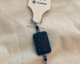 Puede incluir: Un llavero de cuentas con un colgante rectangular con un diseño de plumas de pavo real en tonos azules y turquesas. El llavero incluye un anillo de metal plateado y un adorno de cuentas colgantes. La etiqueta dice "BeadedKink".