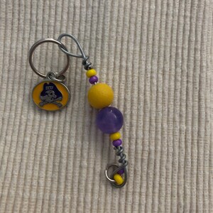Peut inclure: Un porte-clés avec un charm circulaire affichant "ECU" et un logo de tête de mort et d'os croisés en jaune, violet et noir. Il comprend un accent de perles jaunes et violettes.