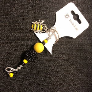 Puede incluir: Un llavero hecho a mano con un dije de abeja plateada, cuentas amarillas y negras y un diseño de alambre plateado. El dije de abeja tiene detalles amarillos y negros. La etiqueta dice "BeadedKink".