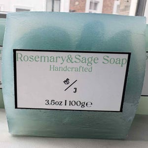 Può includere: Una saponetta rettangolare blu chiaro al rosmarino e salvia. L'involucro trasparente rivela un'etichetta bianca con la scritta "Rosemary & Sage Soap Handcrafted", un'immagine e il peso: 100g.