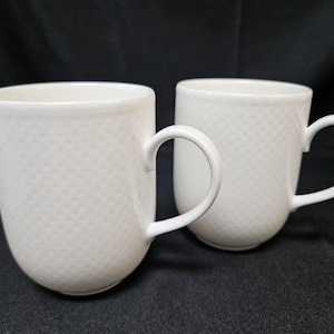 May include: Two white ceramic mugs featuring a subtle, raised dot texture across the exterior surface. Each mug has a smooth, rounded body and a curved handle extending from the side, providing a clean and uniform appearance for these drinkware items.