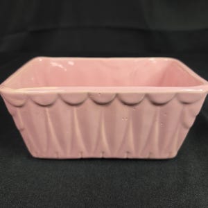 May include: A rectangular ceramic bowl with a glossy light pink finish. The exterior features a decorative scalloped top edge that transitions into vertical, tapered indentations along the sides, creating a textured, pleated appearance.