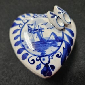 May include: A heart-shaped ceramic trinket box with a white base and blue hand-painted details. The lid features a central windmill illustration surrounded by floral motifs and two small, three-dimensional ceramic clogs attached to the top.