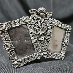 May include: A silver-toned metal double picture frame featuring an ornate design with floral motifs, scrollwork, and a cherub figure at the top center. The frame has two rectangular openings for photographs, surrounded by intricate, raised metal detailing.