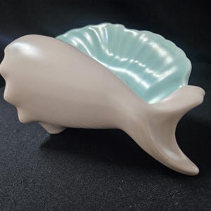 May include: A ceramic dish shaped like a whale with a smooth matte light gray exterior and a glossy light blue interior featuring a ribbed shell-like texture. The whale figure is positioned horizontally with its tail fin extending to the right.